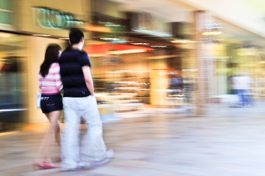 Das Bild zeigt ein Paar, das sich vor Einkaufsläden bewegt. Der Hintergrund ist verschwommen und verdeutlicht Schnelllebigkeit.